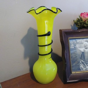 May include: A yellow glass vase with a ruffled top and black swirl design. The vase is sitting on a wooden surface.
