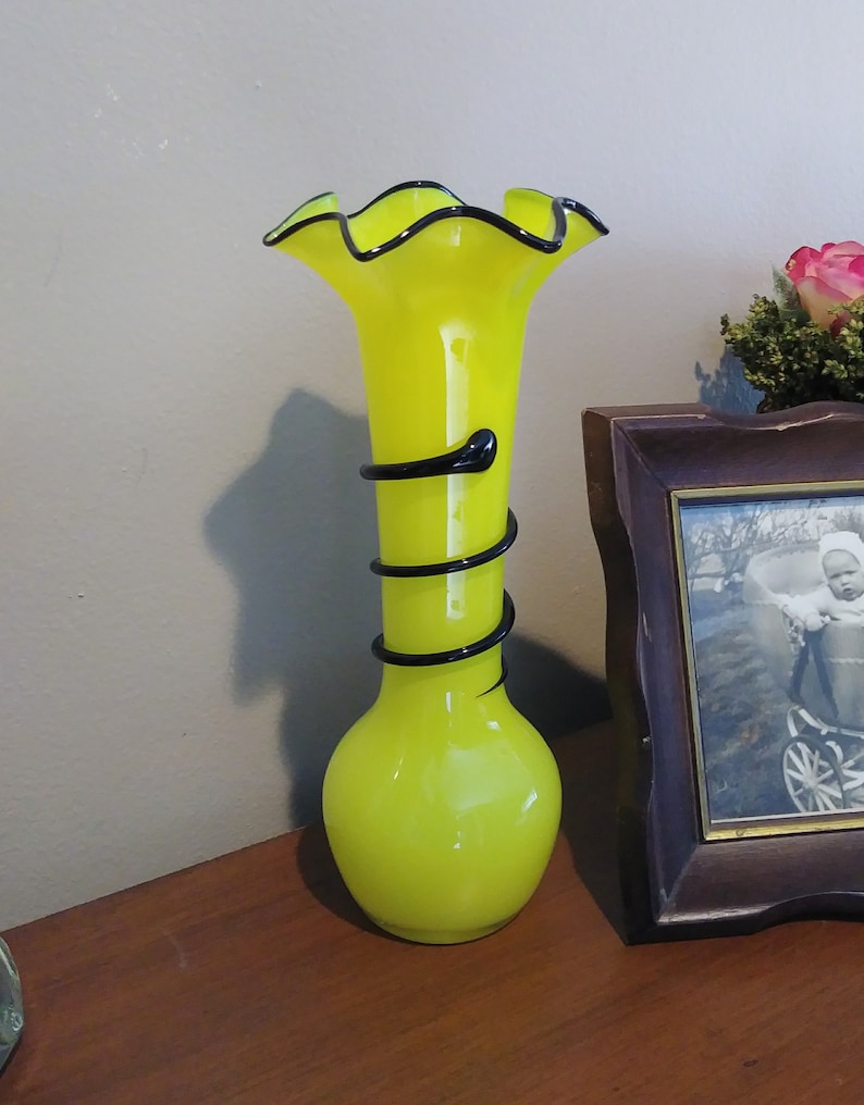 May include: A yellow glass vase with a ruffled top and black swirl design. The vase is sitting on a wooden surface.