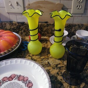 May include: Two yellow glass vases with black accents and ruffled tops. The vases are sitting on a brown countertop.