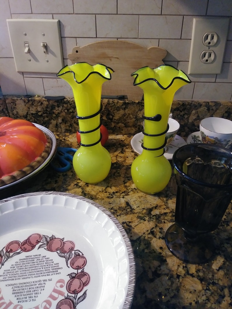 May include: Two yellow glass vases with black accents and ruffled tops. The vases are sitting on a brown countertop.