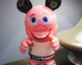 May include: Pink figurine with large ears, wearing sunglasses and a red, white, and blue swimsuit. The ears have firework designs on them.