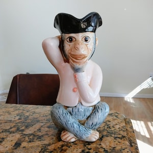 May include: A ceramic monkey figurine, seated with one hand to its head. The monkey wears a black hat, a pink jacket, and blue pants. The figurine has a detailed face and is posed on a speckled countertop.