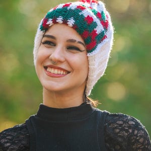 May include: A white knit beanie with a geometric pattern in red and green. The hat is worn by a person wearing a black turtleneck sweater. The background is blurred green.