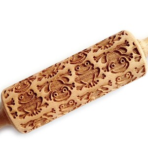 Pode incluir: Um rolo de massa de madeira com um design de sapos esculpidos. O rolo é castanho e tem um acabamento de madeira natural.