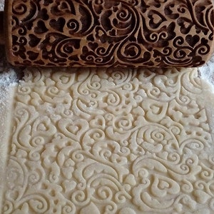 Può includere: Un mattarello di legno con un design intagliato a forma di cuori e volute viene utilizzato per stendere la pasta, creando un motivo in rilievo sulla superficie.