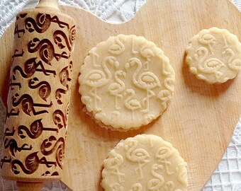 May include: A wooden rolling pin with a flamingo design is used to create cookies with the same design. Three cookies are shown on a wooden cutting board.