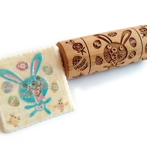 May include: A wooden rolling pin with an engraved design of a blue bunny and Easter eggs. The rolling pin is used to create an imprint of the design on cookie dough.
