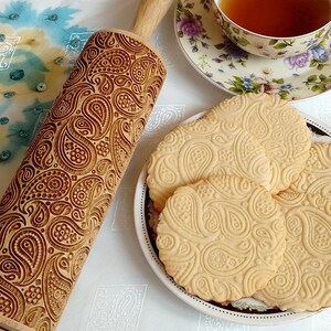 May include: A wooden rolling pin with a paisley pattern is used to create cookies with the same design. The cookies are on a white plate with a black and white border.