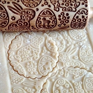 May include: A wooden rolling pin with an engraved Easter design of bunnies and eggs. The rolling pin is being used to roll out dough for cookies. The dough is imprinted with the design from the rolling pin.
