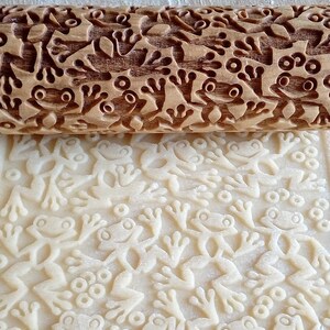 Pode incluir: Um rolo de massa de madeira com um design de sapo é usado para criar um padrão em relevo em uma folha de massa. O rolo tem um design de sapo detalhado esculpido na madeira. A massa é estampada com o design de sapo.