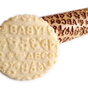 May include: A wooden rolling pin with a baby-themed design. The rolling pin is engraved with letters, numbers, stars, teddy bears, and other baby-related images. The rolling pin is shown rolling out dough, which is imprinted with the design.