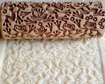 May include: A wooden rolling pin with a frog design is used to create an embossed pattern on a sheet of dough. The rolling pin has a detailed frog design carved into the wood. The dough is imprinted with the frog design.