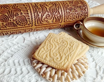 May include: Two square cookies with a seahorse design, made with a wooden rolling pin with a seahorse design. The cookies are on a white crocheted surface with seashells.