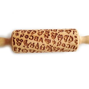 May include: A wooden rolling pin with a pattern of baby-related icons, including footprints, stars, a pacifier, a rattle, a stroller, and the letters ABC. The rolling pin is made of natural wood and has a light brown color.