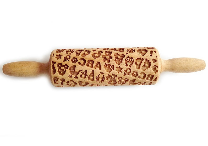 May include: A wooden rolling pin with a pattern of baby-related icons, including footprints, stars, a pacifier, a rattle, a stroller, and the letters ABC. The rolling pin is made of natural wood and has a light brown color.