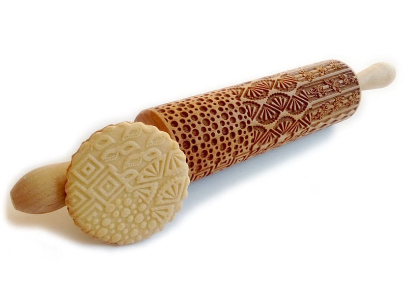 May include: A wooden rolling pin with a decorative pattern carved into the surface. The pattern includes circles, squares, and leaves. The rolling pin is shown with a cookie that has been rolled with the pin, showing the pattern imprinted on the dough.