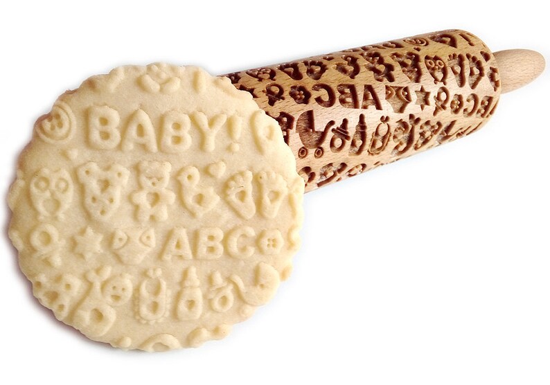 May include: A wooden rolling pin with a baby-themed design. The rolling pin is engraved with letters, numbers, stars, teddy bears, and other baby-related images. The rolling pin is shown rolling out dough, which is imprinted with the design.