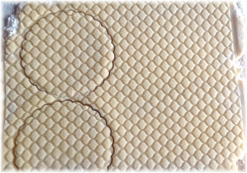 Puede incluir: Primer plano de una l&aacute;mina de masa con un patr&oacute;n de diamantes y dos recortes circulares con bordes festoneados. La masa es de color beige claro y el patr&oacute;n est&aacute; grabado.