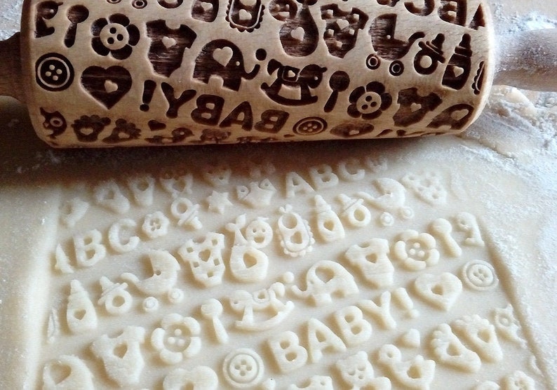 May include: A wooden rolling pin with a baby-themed design, including letters, animals, and other baby-related symbols. The rolling pin is used to create impressions in dough, such as for cookies or fondant.