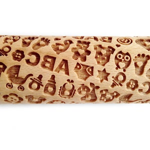 May include: A wooden rolling pin with a baby-themed design. The rolling pin is covered in various baby-related images, including a pacifier, a baby bottle, a rattle, a baby carriage, a teddy bear, a star, a heart, and the letters A, B, and C.