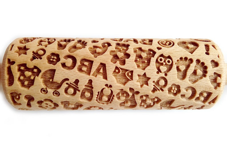 May include: A wooden rolling pin with a baby-themed design. The rolling pin is covered in various baby-related images, including a pacifier, a baby bottle, a rattle, a baby carriage, a teddy bear, a star, a heart, and the letters A, B, and C.