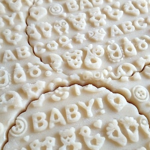 May include: Close-up of a batch of unbaked sugar cookies with various baby-themed designs, including letters, animals, and the word "BABY!"