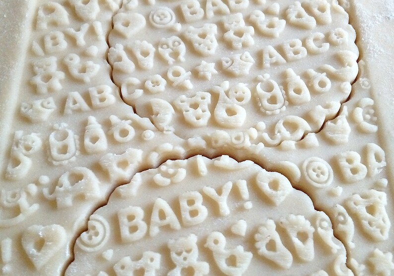 May include: Close-up of a batch of unbaked sugar cookies with various baby-themed designs, including letters, animals, and the word "BABY!"