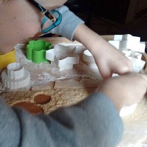 May include: A person using various cookie cutters to shape dough on a wooden surface. The cutters are in the shapes of a flower, star, and circle. Two patterned rolling pins are also visible.