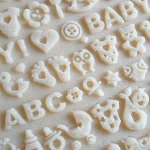 May include: A close-up of a white surface with various cookie cutters pressed into the dough. The cutters include letters, numbers, and baby-themed shapes like a bird, a teddy bear, a pacifier, and a baby onesie.