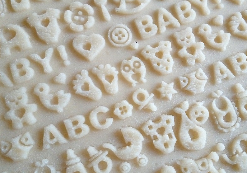 May include: A close-up of a white surface with various cookie cutters pressed into the dough. The cutters include letters, numbers, and baby-themed shapes like a bird, a teddy bear, a pacifier, and a baby onesie.