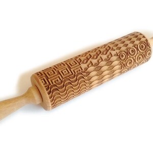 May include: A wooden rolling pin with a carved design of geometric patterns, waves, and circles. The rolling pin is made of light brown wood and has a handle on one end.