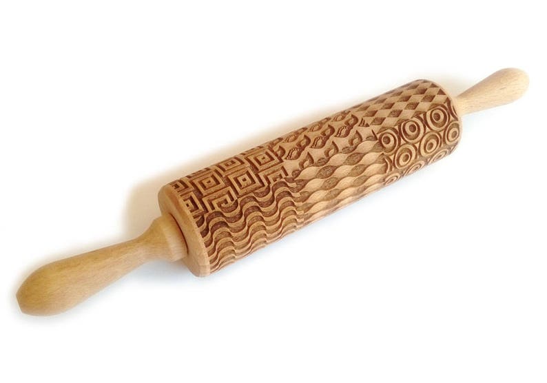 May include: A wooden rolling pin with a carved design of geometric patterns, waves, and circles. The rolling pin is made of light brown wood and has a handle on one end.