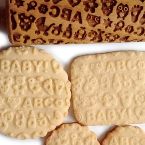 May include: A wooden rolling pin with a baby-themed design, including letters, animals, and other symbols. The rolling pin is used to create decorative patterns on cookies. Four cookies are shown with the same design, including the words "BABY!" and "ABC".