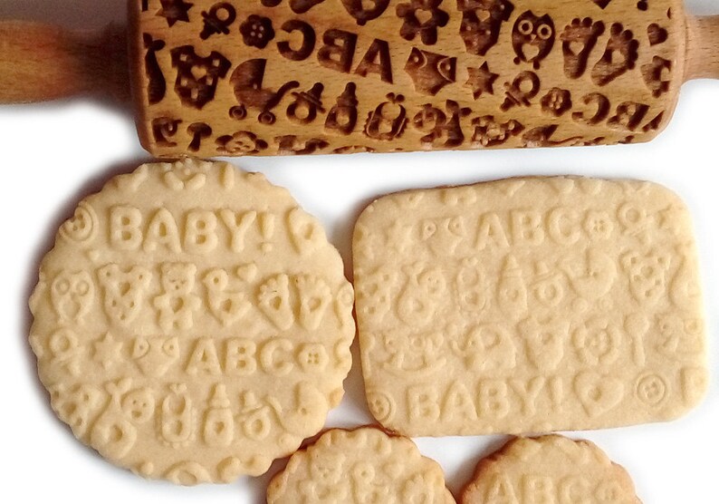 May include: A wooden rolling pin with a baby-themed design, including letters, animals, and other symbols. The rolling pin is used to create decorative patterns on cookies. Four cookies are shown with the same design, including the words "BABY!" and "ABC".
