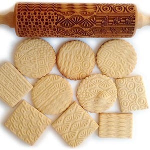 May include: A wooden rolling pin with various engraved patterns, along with several cookies imprinted with the same patterns. The cookies are light brown and have a slightly textured surface.