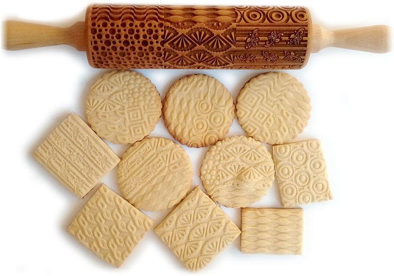 May include: A wooden rolling pin with various engraved patterns, along with several cookies imprinted with the same patterns. The cookies are light brown and have a slightly textured surface.