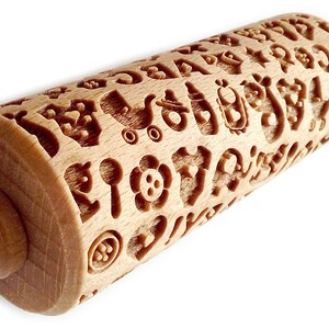 May include: A wooden rolling pin with a carved design of various baby-themed symbols, including a pacifier, a rattle, a baby carriage, and a heart. The rolling pin is made of light brown wood and has a smooth, polished finish.