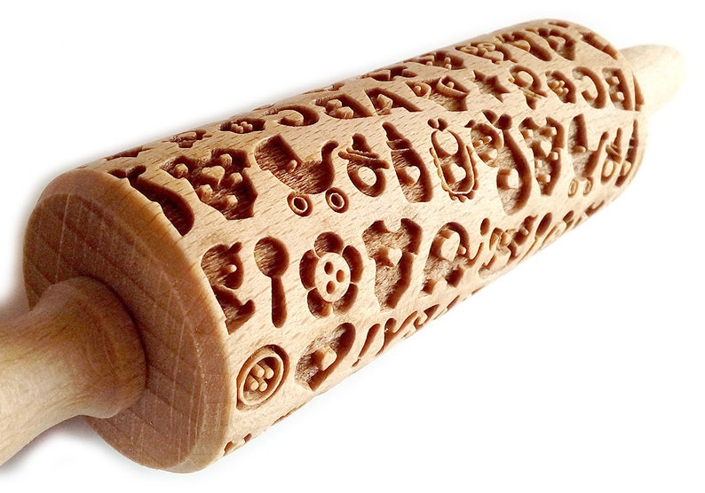 May include: A wooden rolling pin with a carved design of various baby-themed symbols, including a pacifier, a rattle, a baby carriage, and a heart. The rolling pin is made of light brown wood and has a smooth, polished finish.