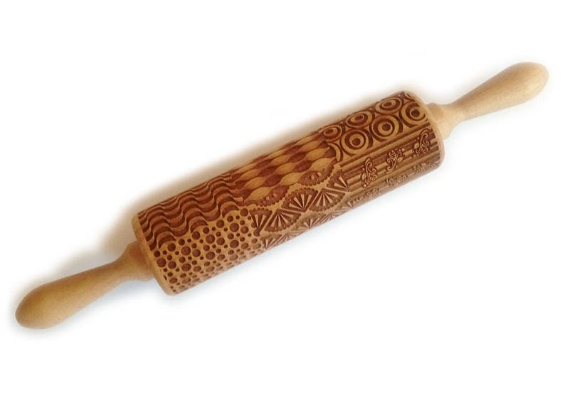 May include: A wooden rolling pin with a decorative engraved pattern of circles, waves, and geometric shapes. The rolling pin is brown and has two wooden handles.
