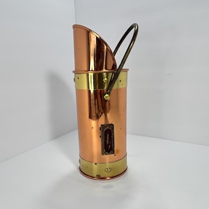 May include: A copper and brass coal scuttle with a curved top and a handle. The cylindrical body features brass bands at the top and bottom, and a small rectangular opening. The handle is arched and made of brass.