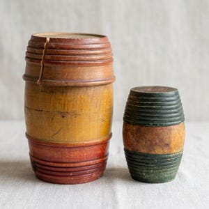 May include: Two cylindrical wooden containers. The larger container features a light brown center with dark red top and bottom sections, showing a vertical crack. The smaller container has a natural wood center with dark green ends.