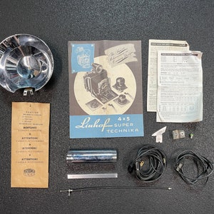 May include: A set of camera accessories for a Linhof 4x5 Super Technika camera. The set includes a flash reflector, a flash cord, a cable release, a lens shade, a manual, and other accessories.