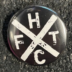 May include: A black button with white text forming an X shape. The letters F, H, T, and C are visible. The button is round and has a glossy finish.