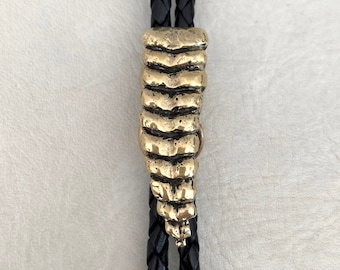 Op de afbeelding: Een bolo-das met een gestructureerde, goudkleurige metalen schuif in de vorm van een ratelslangratel. De schuif is geplaatst op twee zwarte gevlochten leren koorden die eindigen in gepolijste goudkleurige metalen uiteinden onderaan het zichtbare deel.