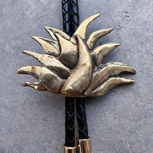 May include: A gold-toned bolo tie with a stylized, abstract floral design. The tie is made of braided black leather cord.