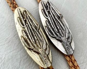 Op de afbeelding: Twee bolo-strikken met zilveren en gouden metalen uiteinden met een gedetailleerd ontwerp van een cactusplant. De bolo-strikken zijn gemaakt van bruin lederen koord.