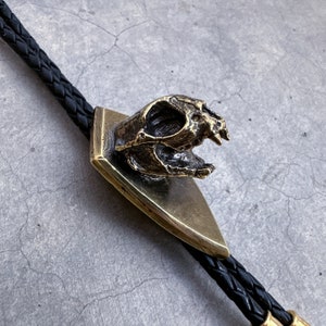May include: A brass bolo tie featuring a detailed skull design. The skull is mounted on a triangular base and has a textured finish. The bolo tie is made with black braided leather cord.