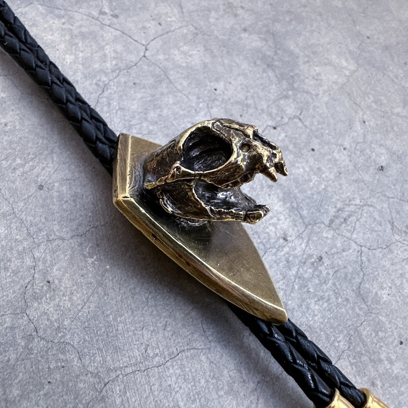 May include: A brass bolo tie featuring a detailed skull design. The skull is mounted on a triangular base and has a textured finish. The bolo tie is made with black braided leather cord.