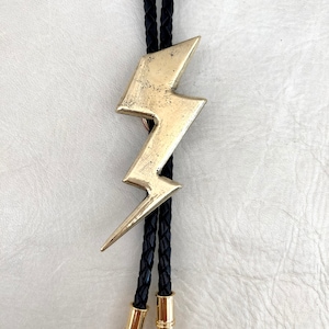 Op de afbeelding: Een bolo-das met een goudkleurige metalen hanger in de vorm van een bliksemschicht met een verweerde, getextureerde afwerking. De hanger is bevestigd aan een zwart gevlochten leren koord dat eindigt in twee gepolijste goudkleurige metalen uiteinden.