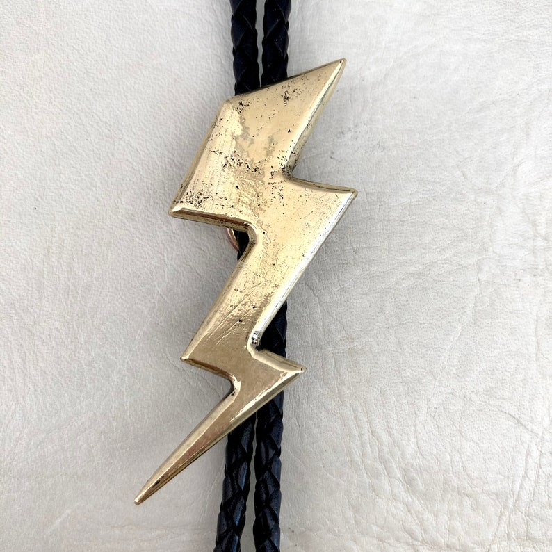 Op de afbeelding: Een bolo-das met een goudkleurige metalen hanger in de vorm van een bliksemschicht met een verweerde, getextureerde afwerking. De hanger is bevestigd aan een zwart gevlochten koord dat verticaal achter de bliksemschichtvorm langs loopt.